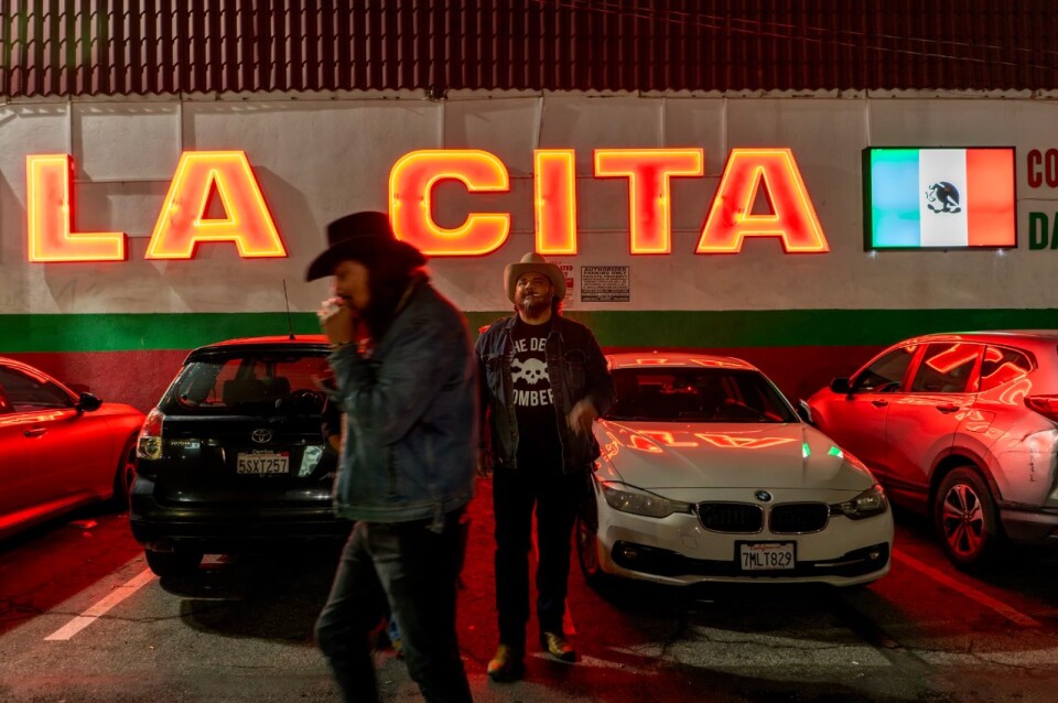 Two men stand in a parking lot at night with a large bright sign on a building behind them. The sign reads "LA CITA" and there is also a lighted Mexican flag.