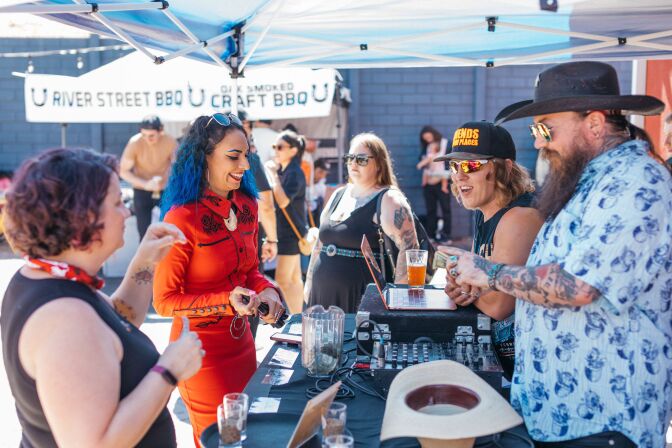 A woman with medium skin tone and blue hair smiles facing a table with DJ equipment and two people with light skin tone wearing hats and sunglasses.