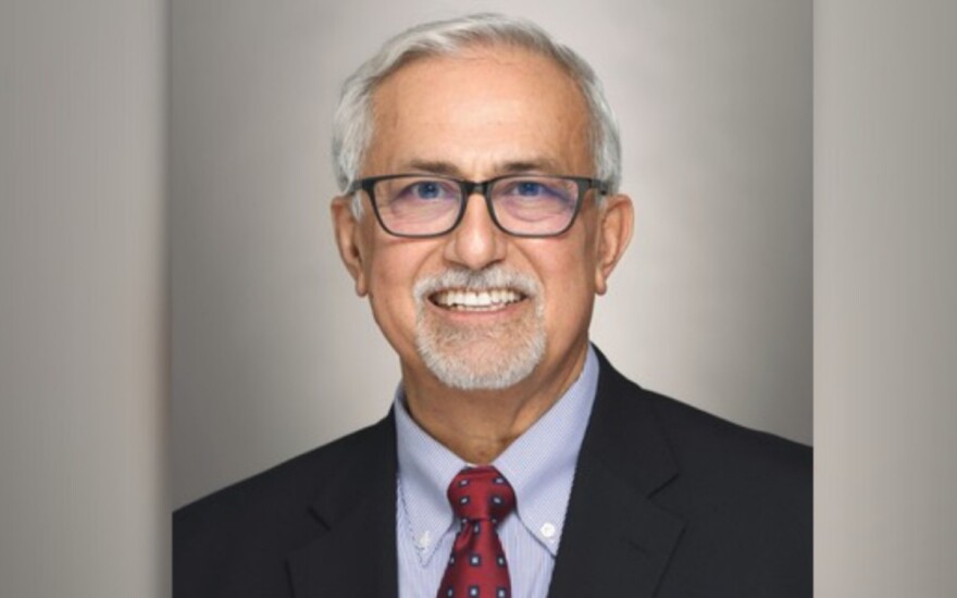A headshot of a man who appears to be white or Latino. He is smiling and looking at the camera. He wears glasses and has a grey goatee and hair. He wears a dark suit with a light blue shirt and red tie with white dots. 