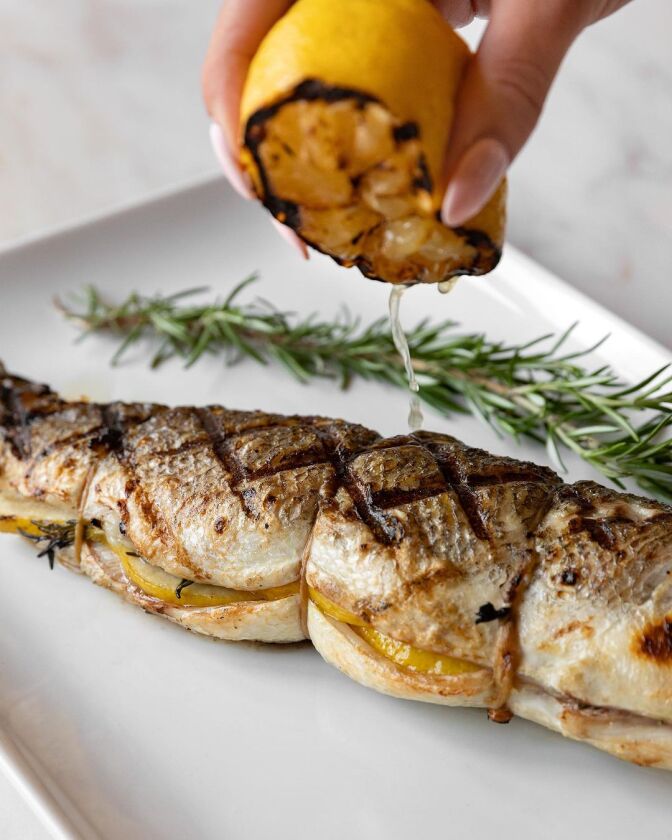 A cooked branzino lies on a white plate, next to a sprig of rosemary. Above it, a manicured hand is squeezing lemon juice onto the fish.
