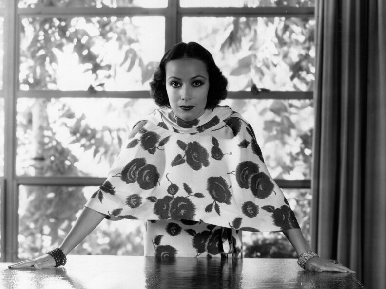 A woman stands in front of a window with both hands on a table. Her outfit has large poppies on it.