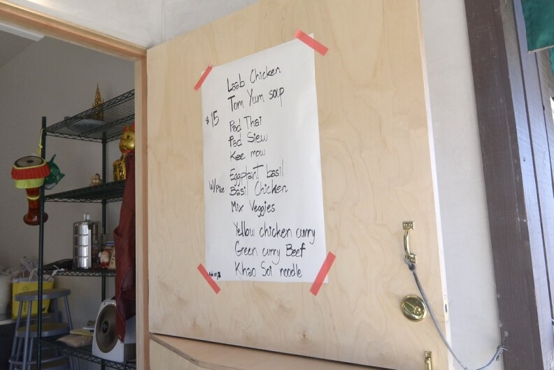 A handwritten menu is displayed on a large piece of white butcher paper. The paper is attached to a light wooden door that stands open, held in place with bright red painter's tape. The menu features various Thai dishes, including pad Thai, green curry with beef, and tom yum soup.