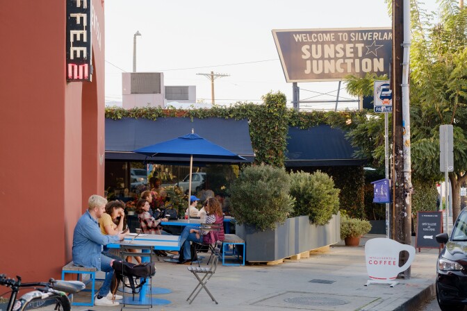 People sit at tables outside Intelligentisa coffee shop in Silver Lake. In the background, a sign reads "Welcome to Silver Lake Sunset Junction."