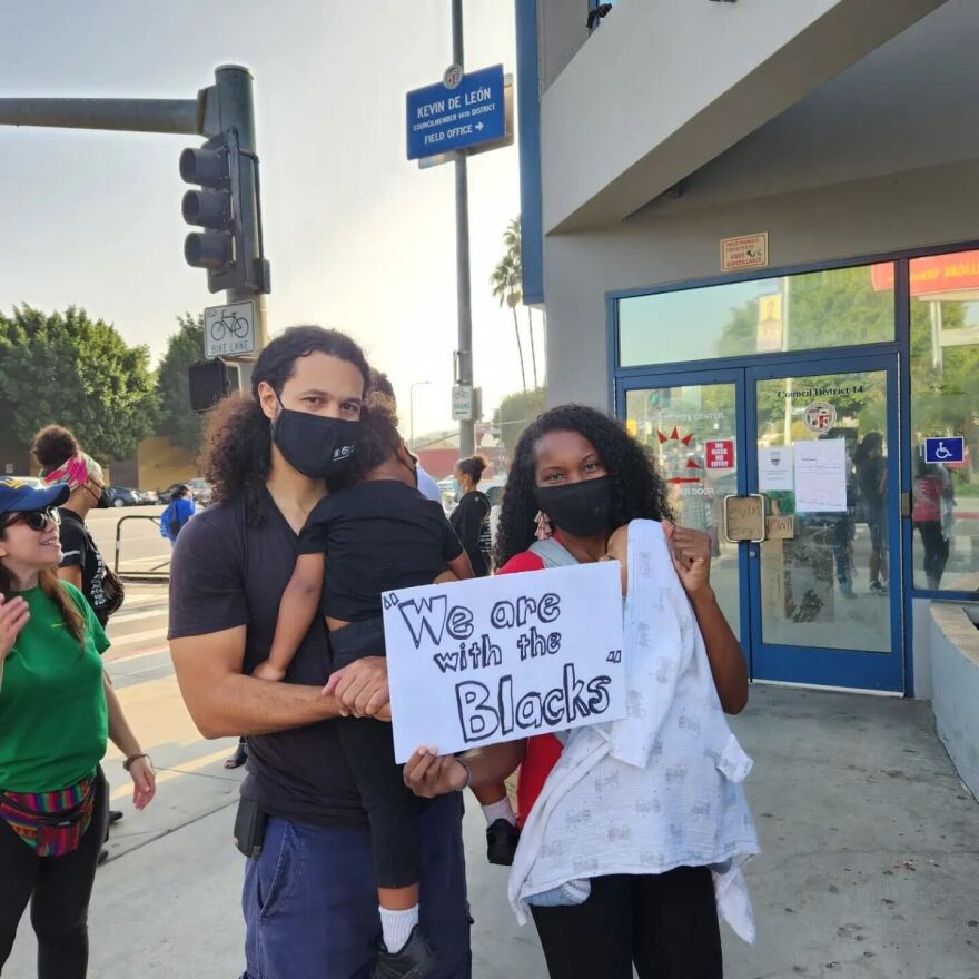 A family poses for a photo on a street corner during a protest. They're standing in front of a grey building with blue piping and doors with windows. A dark-skinned Black woman with long curly black hair wears a black mask over her mouth and nose, a red short sleeve shirt and black pants, and holds her light-skinned brown baby, who is covered with a white patterned blanket. She holds a white and black sign that says "We are with the Blacks." Her husband, a light-skinned Black man, stands next to her, he has curly black hair and wears a black mask over his mouth and nose. He is wearing a black short sleeve shirt and blue pants. He is holding their son, a dark-skinned Black child wearing a black shirt, black pants. and white socks and black shoes.