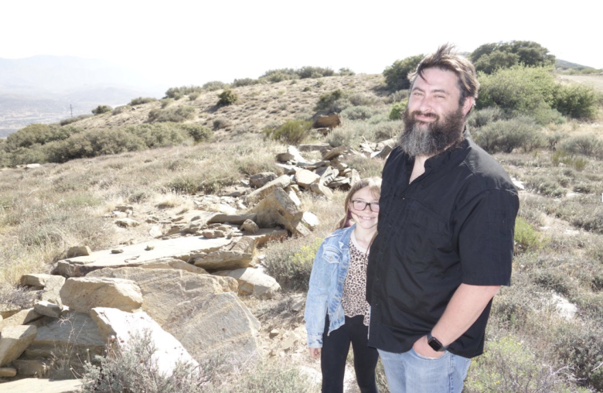 Jeremiah Owen, head of Acton Town Council, standing on a ridge overlooking Acton, with his young daughter, Paige. Jeremiah is wearing a black shirt and blue jeans and has brown hair and a quite long beard and moustache.