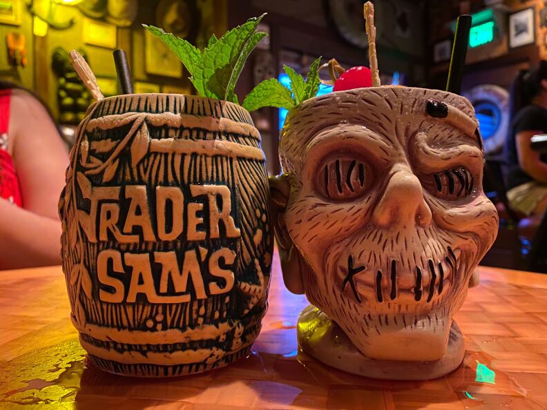 Two tiki mugs sit on a wooden table: One is shaped like a barrel, the other is shaped like a zombie skull.