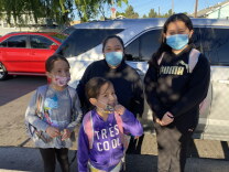 Four individuals — a mother in a black shirt and blue surgical mask, and her three daughters — pose for a photo in front of a car.