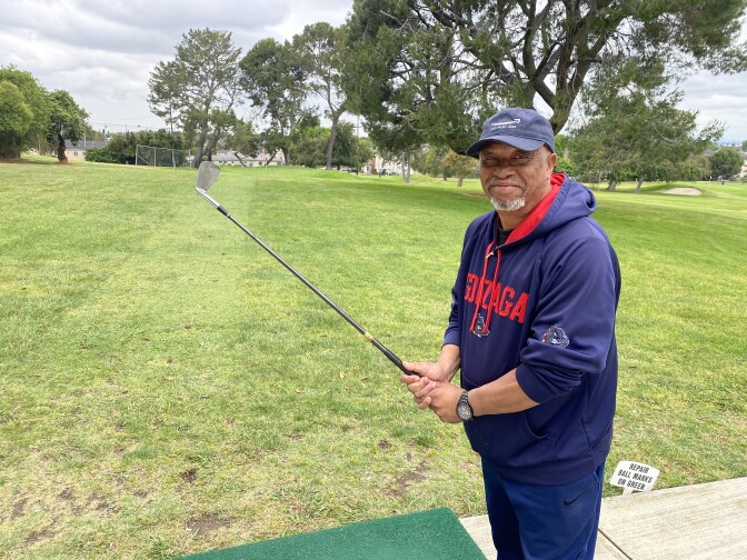 A Black man wearing a blue Gonzaga sweatshirt with "Gonzaga" in red lettering on the front and with a light blue baseball cap, looks at the camera while holding a golf club with a golf course behind him.
