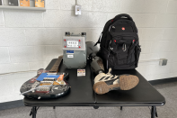 A small black table is seen in a room, there is a white brick wall behind the table and a gray linoleum floor under it. On the table an electric guitar, and electric meter, a stack of baseball cards, a prosthetic leg with an Adidas shoe attached, and a black backpack are depicted.