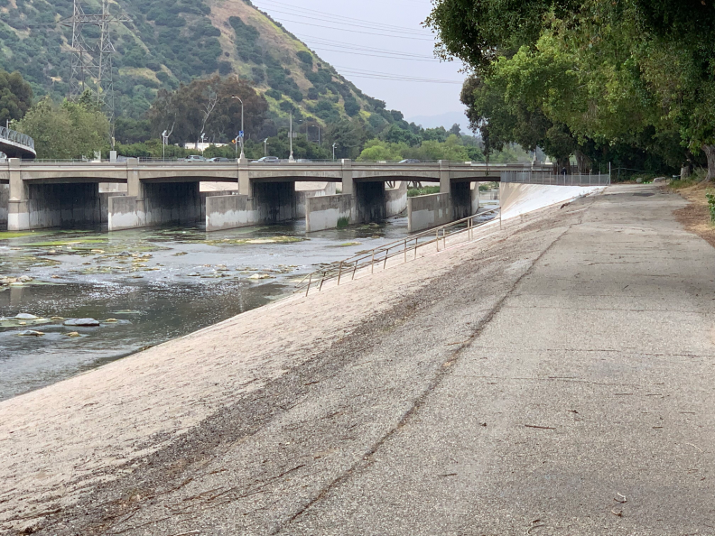 A wide open path leads off into the distance, running along the L.A. River. There is water running in the river. And in the far distance there is a bridge, and signs of busy traffic making its way across the bridge.