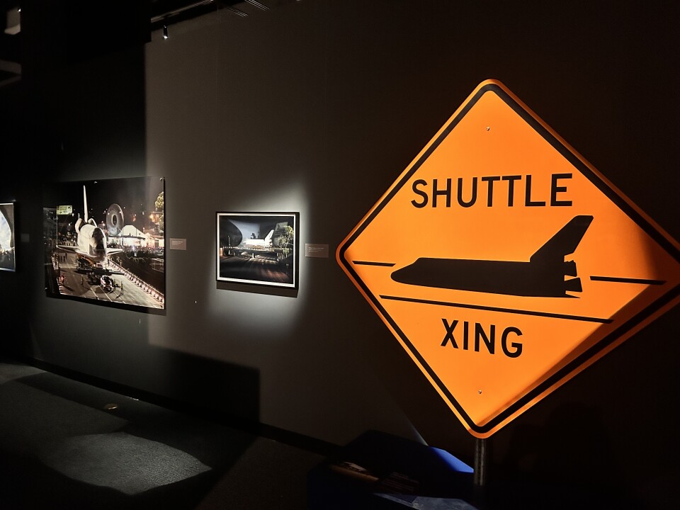 The interior of a museum gallery with several framed photos displayed and well-lit on the wall. A large bright-orange diamond street sign with black text reads "Shuttle Xing"