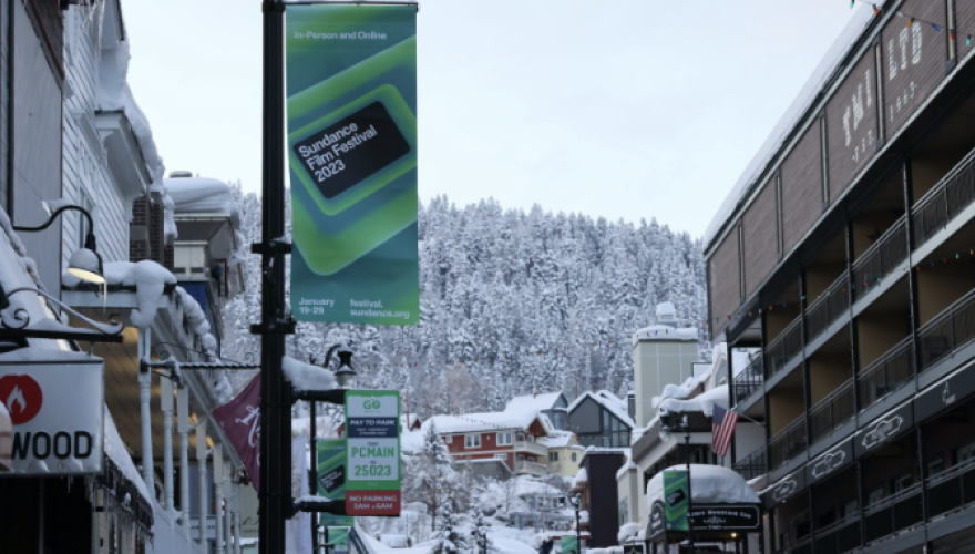 A snowy street scene with snow covered trees in the background, buildings in the foreground with a blue and green sign that says Sundance Film Festival 2023