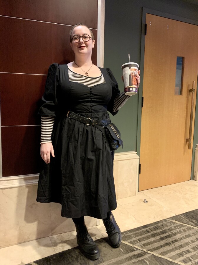 A person with light skin tone and buzz-cut brown hair wears red lipstick, round glasses, a black dress with poofy quarter-length sleeves, a black-and-white striped long-sleeved shirt and black boots. They hold a movie theater drink cup. 