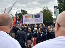 A group of LGBTQ+ supporters stand behind a police line outside the school holding up the sign I hope you know how loved you are in rainbow and trans pride flag colors.