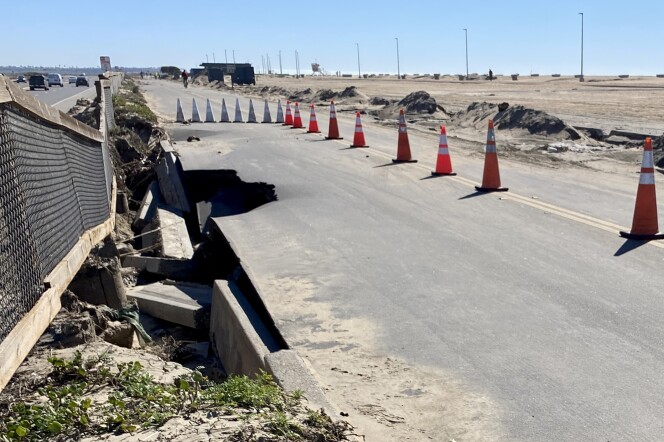 View of a road with the beach on the right and a fence on the left, with a highway on the other side. Orange cones line the road and the pavement is caved in on the left-hand side. 