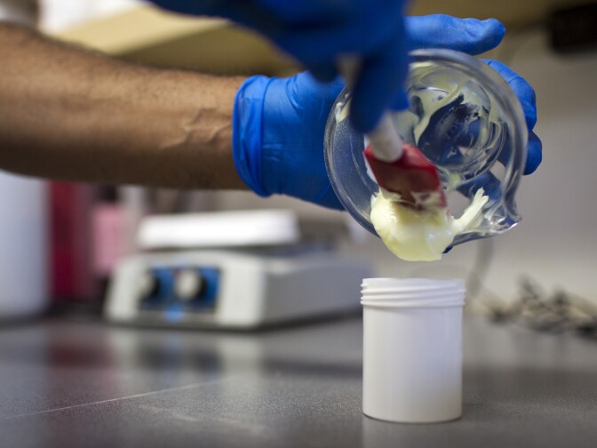 A pharmacist transfers the cream from a mortar to the prescription container.