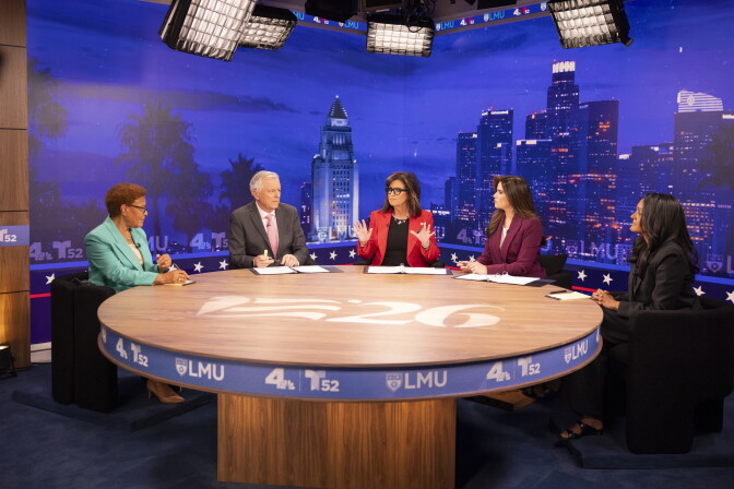 Five people sit at a round table in a TV studio with a Los Angeles skyline backdrop for a debate.