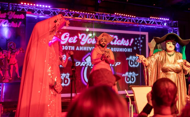 Three drag performers stand on stage they are wearing nun-like costumes. The person on the far right of frame pours what looks like liquor on the stage floor. There's a screen behind them that reads "Get Your Kicks, 30th Anniversary Reunion, Dragstrip 66"