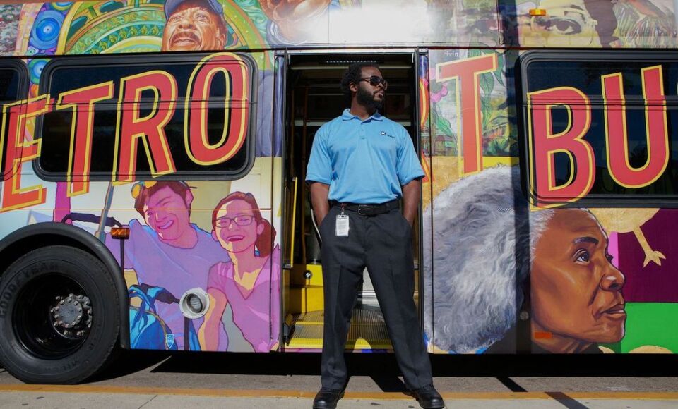 A man wearing a Metro operator uniform poses near the rear exit of the bus. To his right, an artwork depicting two people smiling and looking directly ahead. To his left on the exterior of the bus, an image of an older woman staring into the middle distance.