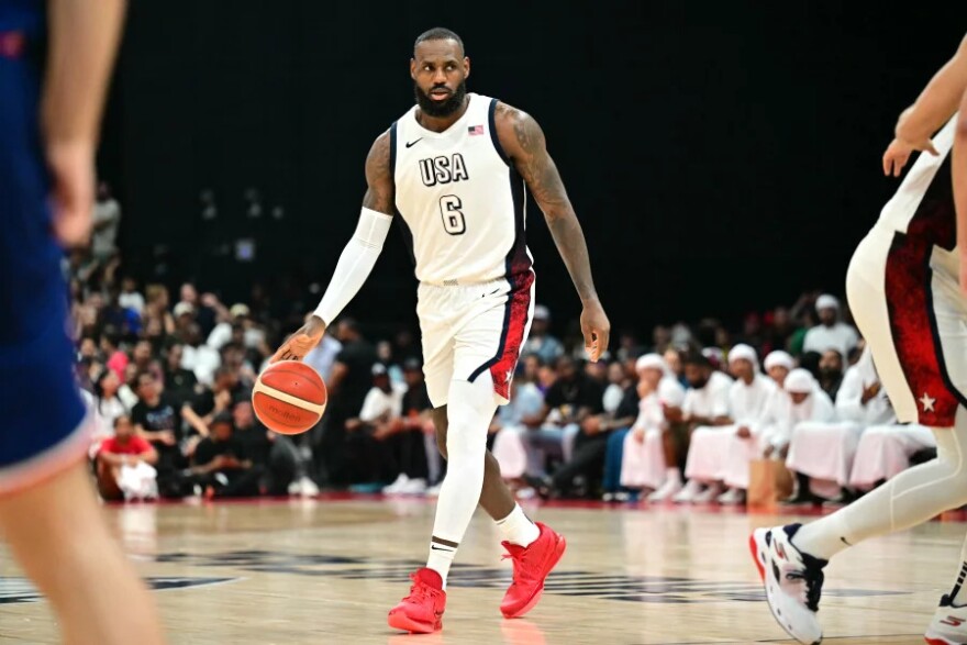 A male presenting basketball player dribbles a basketball on a court with spectators in the background.