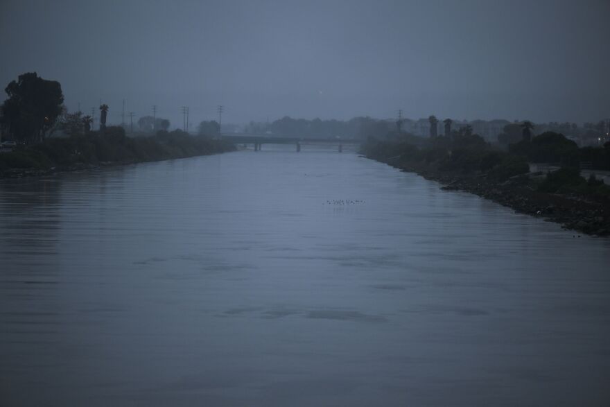 The banks of a creek appear overflowing as rain continues to pour down. A bridge is visible in the distance