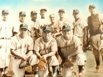 10 men all wearing baseball uniforms stand or kneel. One woman in a dress stands among them.