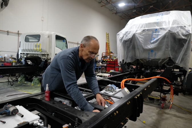 An older man with light-tone skin wears a long-sleeved blue shirt and glasses works on the chassis of a medium-duty truck in a brightly lit warehouse space. 