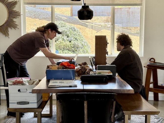 Two men work at a table with recording equipment with a window behind them. 