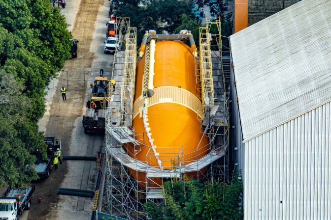 An enormous bright orange tank is surrounded by construction scaffolding. The tank is sitting next to a building outside, dwarfing the construction vehicles and cars parked next to it. 