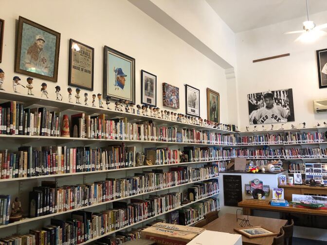 Inside of the Institute of Baseball Studies: four long rows of cataloged books stretch the entity of two walls. There is a small desk with various items on top of it. On top of the shelves are various bobblehead figures of different baseball players. On the wall, above the shelf, are various farmed photographs, artwork, and newspapers on display. In the foreground are desks with various items on top as well. 
