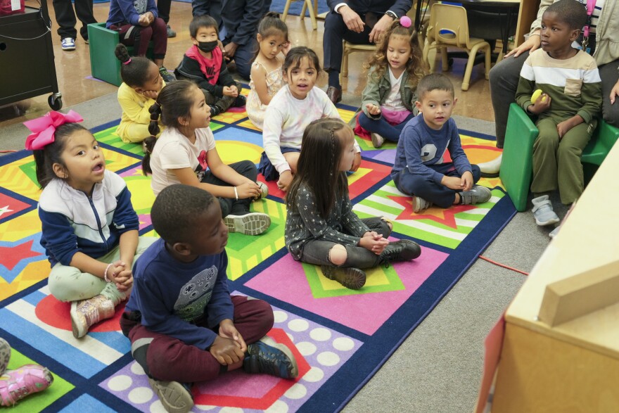 A group of young children with various skin tones sit on a colorful mat inside a classroom. There are some adults sitting near them in the background.