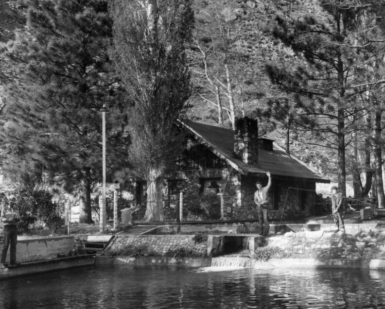 A black-and-white photo of a man standing and waving in front of a cabin set among trees.
