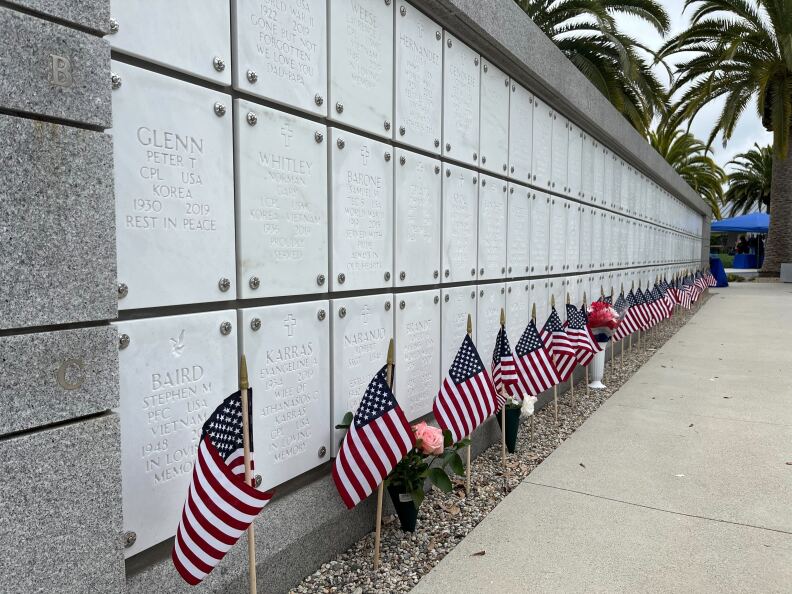 A row of U.S. flags sit in front of a memorial wall of names etched in white stone.