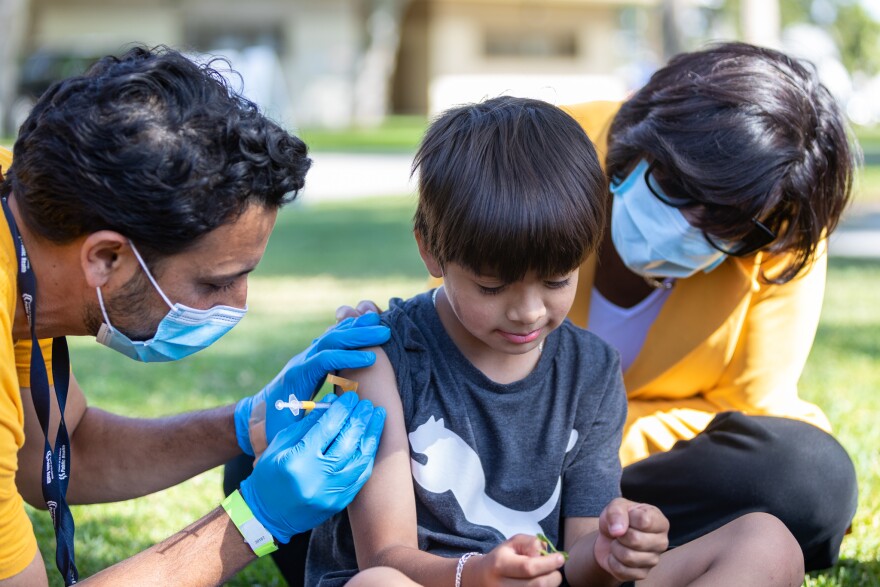 Director of the CDC, Rochelle Walensky (at right) sits on the ground next to a child who is being vaccinated on his right arm by an L.A. County Public Health doctor at Ted Watkins Memorial Park. Walensky and the doctor are wearing protective masks.