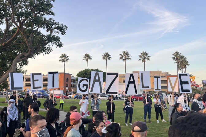 A group of people crowded outside. Some of them are standing and holding signs with one letter each that spells out "Let Gaza Live." 