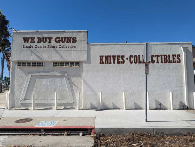 A side picture of a white building in daylight and painted on the building are the words Knives, collectibles, we buy guns in brown.