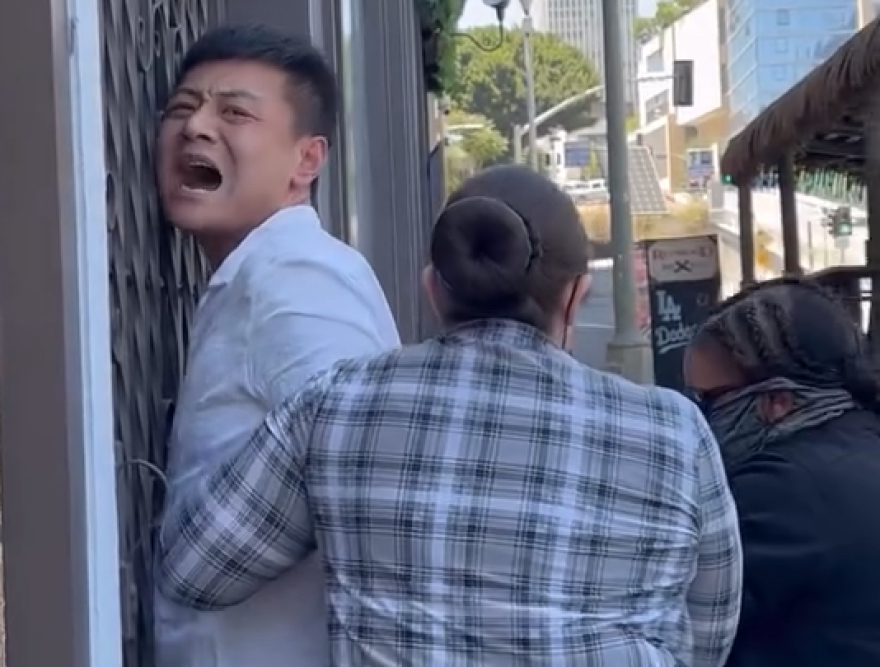 A man is seen yelling for help with his face against a metal door while two women hold his arms.