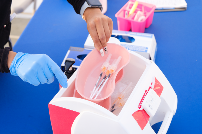 An image of needles of the COVID-19 vaccine in a container. A person, wearing gloves, is holding the container.