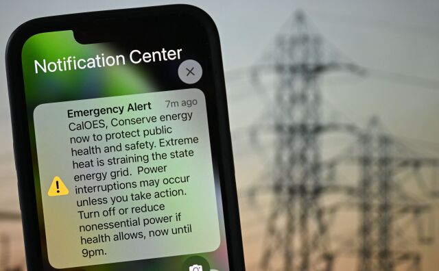 A photo illustration shows a background of electric power infrastructure with an Apple iPhone showing an Emergency Alert notification from CalOES urging the public to conserve energy to protect health and safety.