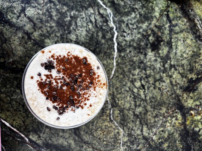 Overhead view of a frothy iced drink topped with a dusting of cocoa powder and crunchy cacao nibs, set on a dark green marble table with white and brown veining.