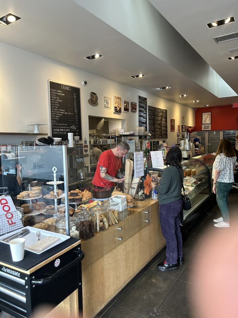The inside of a casual restaurant, with a busy counter and people lining up to give their order to a light skinned man wearing a red t shirt. There are large blackboard signs behind him showing the menu