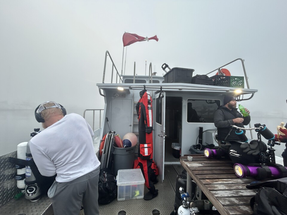 Two men on the deck of a dive boat in foggy weather. The deck has a wooden table covered in diving gear. 