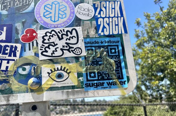 A blue sticker with a qr code and the phrases "Maude Latour," "Sugar Water" and "August 16," plastered on the corner of a street sign and surrounded by other stickers