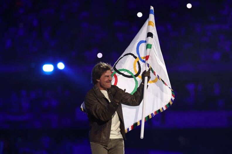 A man with light-tone skin hold the Olympic flag and grins