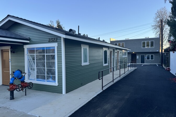 A one-story green building with newly-installed windows sits in the foreground and another two-story complex sits behind. 