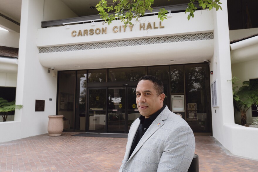 A man with medium skin tone and short hair wearing a gray suit stands in front of a white building with gold text that reads "Carson City Hall."