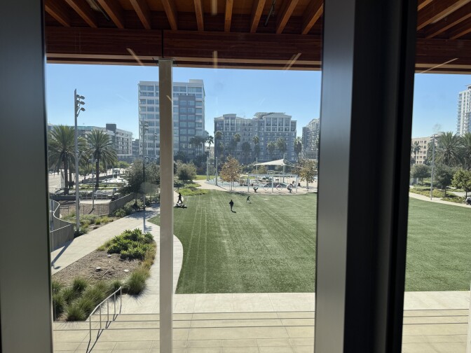 A view through large glass windows looking out onto an open green space in an urban setting.