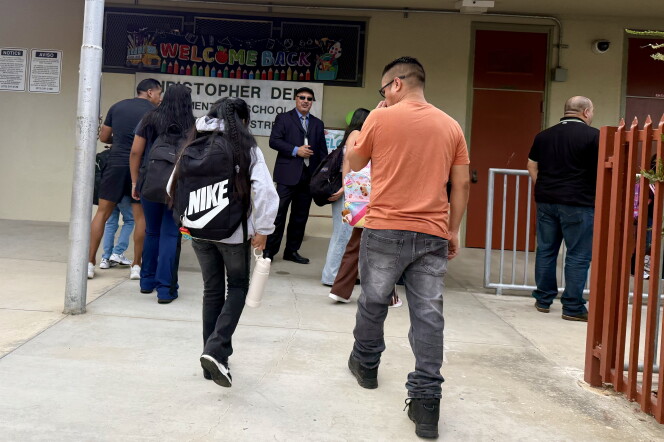 Adults and children wearing backpacks are seen from behind, walking toward the entrance of an elementary school. 