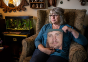 A woman with graying shoulder length care and eyeglasses is seated in a wingback chair holding a large photo of a young man with light skin, green eyes, a mustache and goatee. A fish tank is beside her on a table.