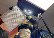 A man in a bright neon yellow safety vest stands on top of a ladder near the ceiling of a hallway while holding a large air filter, perhaps 2 feet long by 2-and-a-half feet wide.